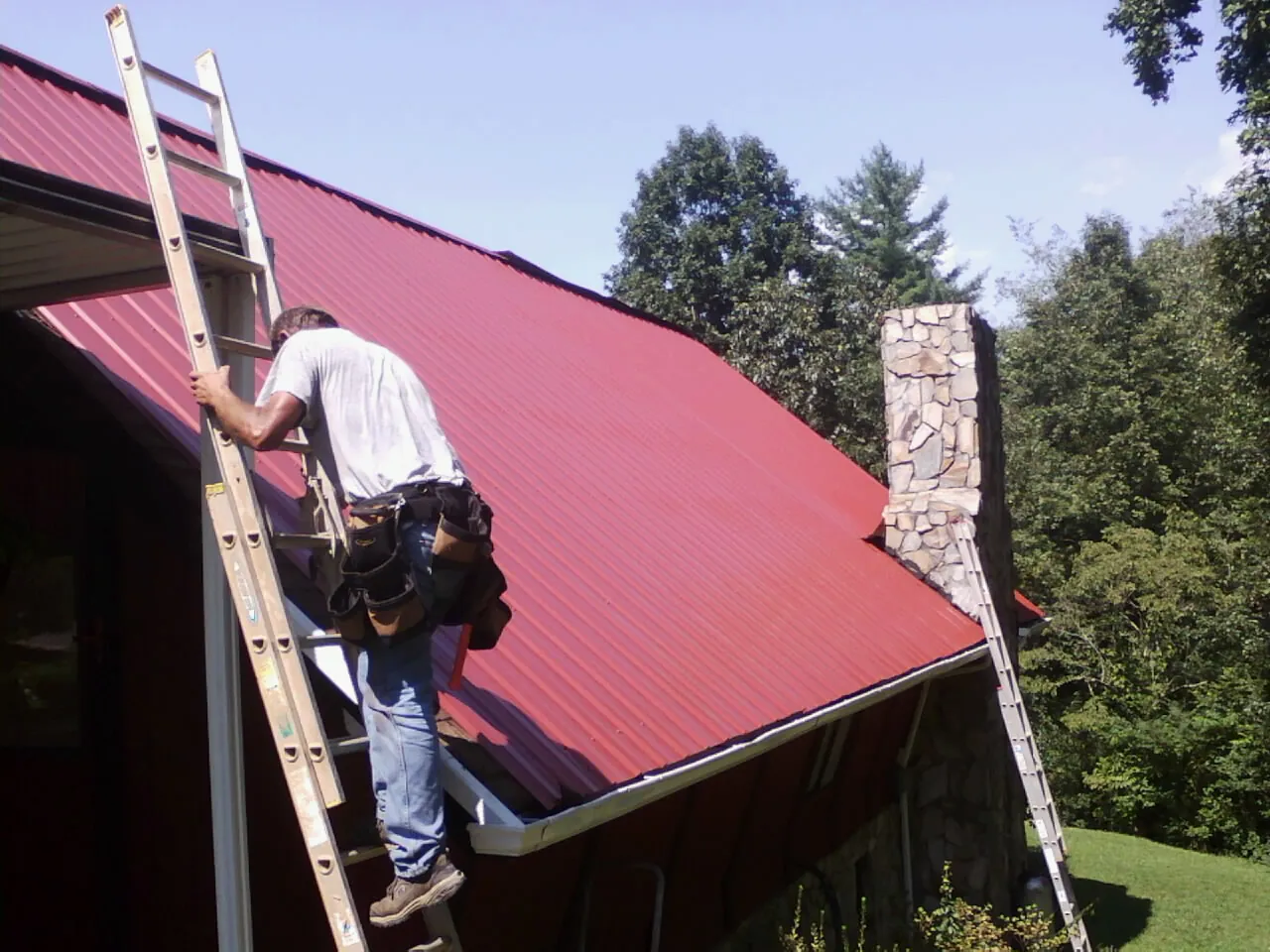 Roofer on a ladder beside a red metal roof and stone chimney — Soffit & Fascia Repair by Roofing Brothers in Cache